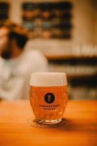 Vrček za pivo, poln z lepo peno. V ozadju oseba, ki sedi v lokalu.